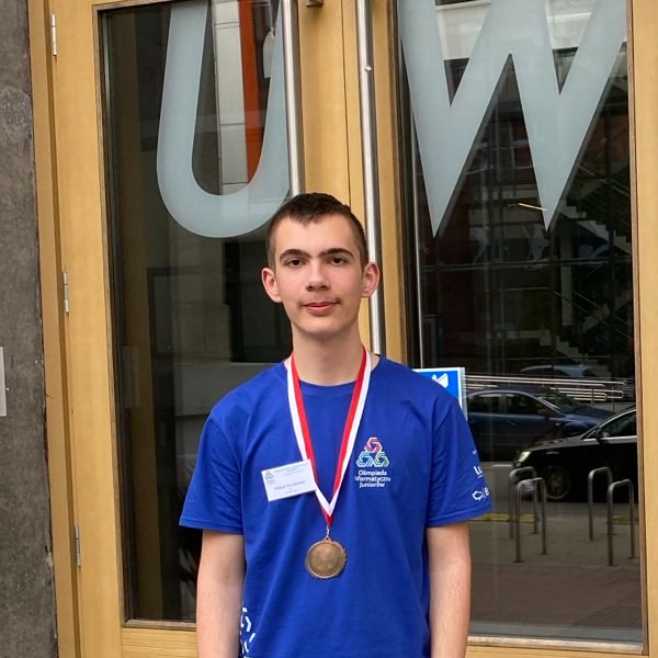 Wiktor Perskawiec smiling and wearing a blue T-shirt with a medal from a competition around his neck. In the background are the doors to the Faculty of Mathematics, Informatics, and Mechanics at the University of Warsaw.